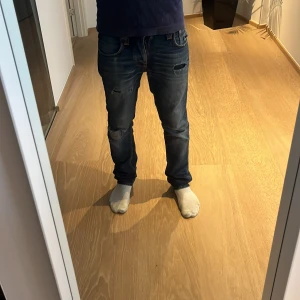 Blå straight jeans från Nudie Jeans - Säljer ett par selvedge straight  jeans från Nudie Jeans med slitna detaljer och hål på knä och fickor. Jeansen har klassisk femficksdesign, orangea sömmar och låg midja. Perfekt för dig som gillar en avslappnad och trendig look.