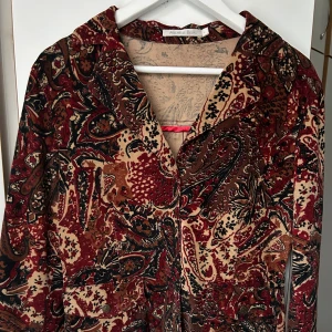 Mönstrad manchesterjacka Michelle Belau - Unik manchesterjacka från Michelle Belau med paisleyinspirerat mönster i rött, brunt, beige och svart. Jackan har klassisk krage, knappar framtill och två fickor. Perfekt för dig som vill sticka ut med en retrovibe och coola detaljer.