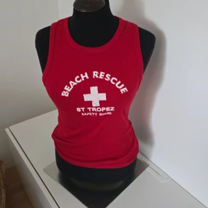Röd topp från F-Spirit, M - Röd topp från F-Spirit med tryck 'BEACH RESCUE ST TROPEZ SAFETY GUARD' och vit korsdetalj framtill.