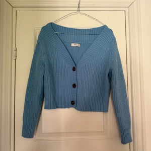 Blå stickad kofta från MNG - Snygg ljusblå stickad kofta från MNG med v-ringad hals och tre svarta knappar framtill. Croppad modell med ribbade muddar och långärmad design. 