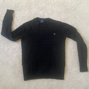 Svart kabelstickad tröja från GANT - Svart kabelstickad tröja från GANT i premium bomull. Klassisk rund halsringning, diskret broderad logga på bröstet och ribbade muddar. Perfekt för dig som gillar stilrena och tidlösa plagg med snygg struktur.