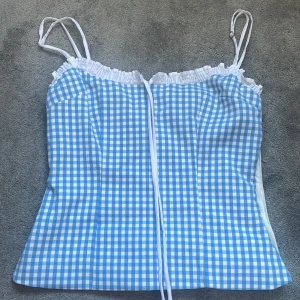 Blåvit rutig korsettopp från Zara - Supersöt korsettopp från Zara i blåvitt rutigt mönster. Toppen har smala, justerbara axelband, rynkad kant upptill och snörning framtill. Perfekt för en somrig look och ger en fräsch vibe. Materialet känns lätt och svalt.