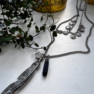Långt lager-på-lager halsband med berlocker - Snyggt lager-på-lager halsband i silverfärgad metall med flera kedjor. Ena kedjan har små runda berlocker med mönster, den längsta har en stor fjäder, en svart droppe och en glittrande sten som hängsmycken. Perfekt statement-piece för att lyfta din outfit. #halsband 