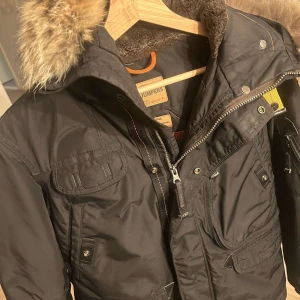 Svart Parajumpers vinterjacka med päls - Svart Parajumpers vinterjacka med flera fickor, gul detalj vid kragen och avtagbar huva med päls. Jackan har quiltat foder, dragkedja och knappar framtill samt klassisk PJS-patch på ärmen. Perfekt för kalla dagar och riktigt snygg streetstil.