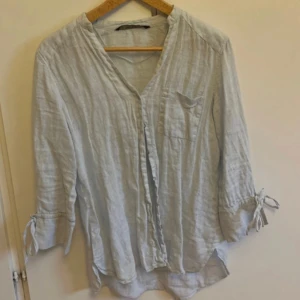 Ljusgrå linneskjorta från Zara  - Luftig ljusgrå skjorta i linne från Zara. Skjortan har v-ringning, knappar framtill, bröstficka och trekvartsärmar med snörning vid ärmsluten. Perfekt för en chill och stilren look under varmare dagar.