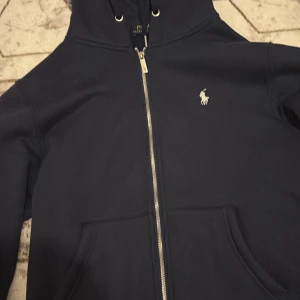Mörkblå hoodie från Polo Ralph Lauren - Mörkblå hoodie från Polo Ralph Lauren med dragkedja, huva och fickor framtill. Klassisk broderad logga på bröstet och mjukt fleecefoder på insidan. Perfekt för dig som gillar stilrena och bekväma plagg. Helt ny säljs endast för 1200kr
