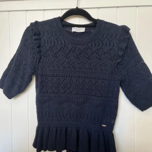 Mörkblå stickad topp Holly & Whyte - Supersöt mörkblå stickad topp från Holly & Whyte med kort ärm, volangdetaljer på axlarna och bred volangkant nertill. Snyggt hålmönster över hela tröjan och rund halsringning. Perfekt för dig som gillar unika detaljer och en feminin vibe.
