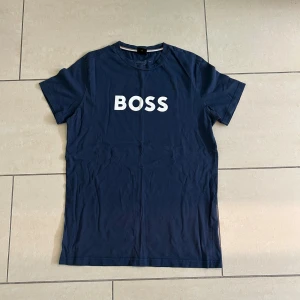 Marinblå BOSS t-shirt i bomull - Snygg marinblå t-shirt från BOSS med vit logga tryckt på bröstet. Klassisk rund hals och korta ärmar. Tillverkad i mjuk bomull för en skön känsla. Perfekt för en clean och stilren look. Knappt använd och säker för bra pris!                                    Pris kan diskuteras!😁