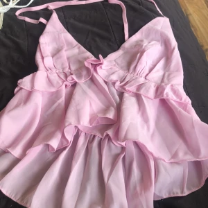 Ljusrosa volangig linnetopp Xs - Supersöt ljusrosa linnetopp med volanger och tunna axelband. Toppen har omlottkänsla framtill och är sydd i ett lätt, transparent syntetmaterial som ger ett flowy intryck. Perfekt för dig som gillar romantisk och feminin stil.