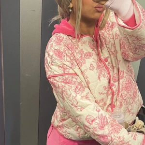 Quiltad blommig jacka i rosa och beige - Superfin quiltad jacka med blommigt mönster i rosa på beige botten. Jackan har rund hals, tryckknappar framtill och rosa kantband. Perfekt för dig som vill sticka ut med en söt och trendig look.