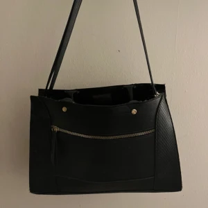 Svart axelväska med dragkedja - Stilren svart axelväska i skinnimitation med raka linjer och guldiga detaljer. Väskan har en praktisk ytterficka med dragkedja och långa axelband. Perfekt storlek för att få plats med allt du behöver till skolan eller stan.