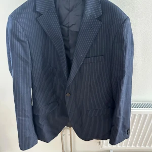 Mörkblå kostym - Snygg mörkblå kostym med tunna vita ränder, består av kavaj och matchande kostymbyxor. Kavajen har klassisk krage, två framfickor med lock och en bröstficka. Byxorna har pressveck och knappstängning. Materialet känns som ullblandning och passformen är slim