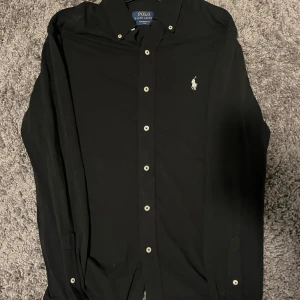 Svart skjorta från Polo Ralph Lauren - Svart skjorta från Polo Ralph Lauren med klassisk krage och vita knappar. Ikoniska Polo-loggan broderad i vitt på bröstet. Skjortan har lång ärm och är gjord i mjuk bomull. Perfekt för dig som gillar stilrena och tidlösa plagg.