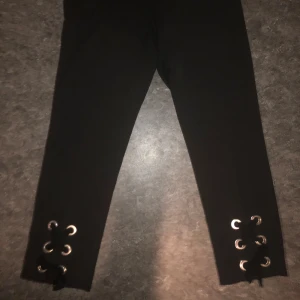 Svarta leggings med snörning nertill - Svarta leggings med snygga snörningsdetaljer och metallöljetter vid bensluten. Tight passform och stretchigt material som sitter skönt. Perfekta för dig som vill ha en stilren look med en cool twist. (Ej rekommenderat för långa kvinnor) 
