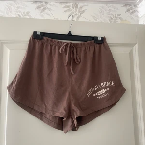 Bruna shorts med Daytona Beach-tryck - Säljer ett par bruna shorts med vitt Daytona Beach-tryck på ena benet. De har elastisk midja med snörning och är tillverkade i mjuk bomull. Modellen är loose fit och passar perfekt för sommardagar eller chill hemma.