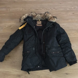 Svart vinterjacka Parajumpers S - Svart vinterjacka från Parajumpers med päls på huvan och snygga gula detaljer. Jackan har flera fickor med dragkedja, ribbade muddar och en klassisk patch på axeln. Perfekt för kalla dagar och har en riktigt schysst streetstil.