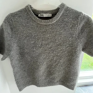 Grå stickad kortärmad tröja Zara - Mysig grå stickad tröja från Zara i storlek S. Tröjan har korta ärmar, rund halsringning och en lite fluffig textur. Perfekt att styla med jeans eller kjol för en chill look.