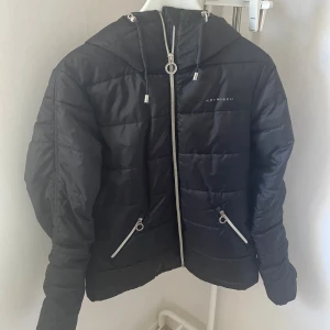 Svart vinterjacka  - Svart vinterjacka från Röhnisch med huva och silvriga dragkedjor. Jackan har quiltad design, två snedställda fickor med dragkedja och snygga detaljer vid snörningen. Storlek 36