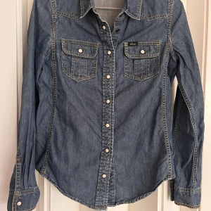 Jeansskjorta från Lee XS - Snygg jeansskjorta från Lee i klassisk blå denim med kontrastsömmar och tryckknappar. Skjortan har två bröstfickor med lock och lång ärm. Perfekt för dig som gillar en tidlös och cool look.
