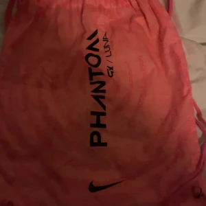 Nike Phantom GX Luna rosa fotbollsskor Elite - Nike Phantom GX Luna fotbollsskor ELITE, men påsen är sönder i smörat derför så billigt pris. De är en snygg rosa färg med svarta detaljer och Nike Swoosh. Skorna har snörning och ovandel i syntetmaterial med textilinslag för bra grepp och känsla på planen. Sulan är designad för optimalt fäste på gräs. Kommer med matchande rosa påse. 