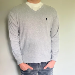 Ralph Lauren tröja  - Snygg grå v-ringad tröja från Polo Ralph Lauren i mjuk pima bomull. Klassisk passform med lång ärm och diskret broderad logga på bröstet. Perfekt till jeans eller skjorta för en clean och stilren look.