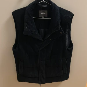 Svart manchesterväst från Dressmann - Säljer en svart manchesterväst från Dressmann i storlek M. Västen har hög krage, dragkedja och tryckknappar framtill samt praktiska fickor. Klassisk och stilren modell i mjukt manchestertyg, perfekt för lager-på-lager-stil.