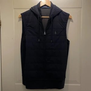 Mörkblå väst från Polo Ralph Lauren - Marinblå quiltad väst från Polo Ralph Lauren med huva och dragkedja framtill. Västen har broderad logga på bröstet och justerbara snören vid huvan. Perfekt för lager-på-lager och stilren look. Tunn väst som är perfekt som mellanlager! Storlek S. Kontakta mig vid minsta fråga eller fundering! Priset kan diskuteras! 