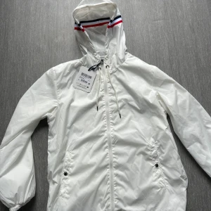 Vit vindjacka från Moncler med huva - Vit vindjacka från Moncler med huva och dragkedja framtill. Jackan har Moncler-logga på ärmen och snygga ränder i rött, vitt och blått på huvkanten. Två fickor med knappstängning och justerbar dragsko vid huvan. Perfekt för dig som vill ha en clean och sportig look.