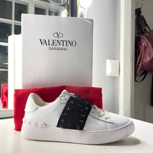 Valentino Rockstud vita sneakers - Snygga vita Valentino Garavani Rockstud sneakers med svart dekorerad rem över vristen, prydd med nitar och pärlor. Skorna har rund tå, snörning och är tillverkade i skinn. Perfekt för dig som vill sticka ut med en lyxig och edgy look.