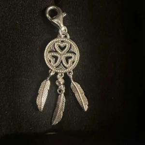 Thomas Sabo berlock - Unik silverfärgad berlock med rund form, tre hängande fjädrar och detaljerade hjärtmönster i mitten. Perfekt att fästa på halsband eller armband för en personlig touch.