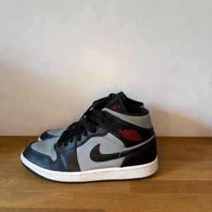 Nike Air Jordan 1 Mid svart/grå - Nike Air Jordan 1 Mid i svart och grått skinn med röda detaljer. Klassisk siluett med svart swoosh, perforerad tå och snörning. Mellanhög modell med vit sula och svart yttersula. Röd Jumpman-logga på plösen och Air Jordan Wings på sidan.