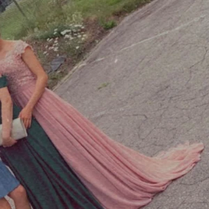 Rosa långklänning med spets och släp - Magisk rosa långklänning med spets upptill och vackert släp. Klänningen har v-ringad hals, korta ärmar och omlottkänsla i kjolen. Perfekt för bal eller student. Materialet känns lyxigt och faller snyggt.