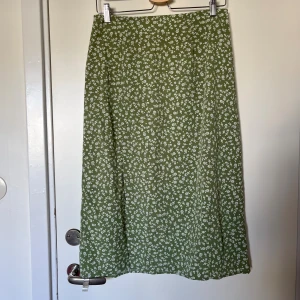 Grön blommig midikjol - Snygg grön midikjol med vitt blommönster. Kjolen har en rak passform och hög midja, dragkedja i sidan. Liten slits. Lätt material. Köpt second hand. 74 cm i midjemått, passar s-m. 