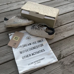 Golden Goose Superstar sneakers vit/grå/blå - ✨✨✨PRIS KAN DISKUTERAS VID EN SNABB AFFÄR ✨✨✨Golden Goose Superstar sneakers i vit skinn med grå snörning och mörk blå häl. Klassisk stjärndetalj på sidan och platt sula med slitna detaljer för en cool look. Kommer med originalpåse och box. De är storlek 42 men är rätt så små i storleken. Så 41 är rätt storlek. Kom privat för mått. Dem är endast använda max 6ggr och säljs för dem var försmå pris kan diskuteras vid en snabb affär 😁