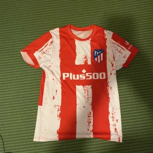 Atlético Madrid João Félix matchtröja - Säljer en Atlético Madrid fotbollströja med João Félix och nummer 7 på ryggen. Tröjan är röd och vit med breda, randiga mönster och klubbmärke på bröstet. Kortärmad modell i lätt och ventilerande material, perfekt för fotboll eller träning.