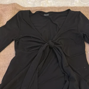 Svart omlottopp från Gina Tricot XS - Svart omlottopp från Gina Tricot i storlek XS. Toppen har lång ärm, djup v-ringning och knyt framtill som ger en snygg och trendig look. Tunt och mjukt material som känns skönt mot huden. Perfekt att styla med jeans eller kjol.