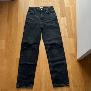 Svarta jeans med hål i knät från Gina Tricot - Svarta jeans från Gina Tricot i rak modell med snygga hål vid knäna. Jeansen har fem fickor, hög midja och klassisk knappgylf. 