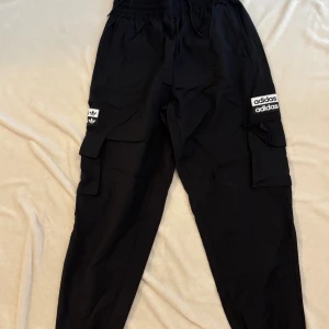 Svarta cargobyxor från Adidas - Svarta cargobyxor från Adidas med stora fickor på sidorna och vita Adidas-loggor på båda benen. Byxorna har elastisk midja med snörning och är tillverkade i ett mjukt och bekvämt material. Perfekta för en sportig och avslappnad stil. Oanvänd.