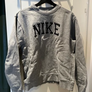 Grå sweatshirt från Nike i medium - Grå sweatshirt från Nike med broderad svart logga och text på bröstet. Klassisk rund halsringning, ribbade muddar och mjukt fleecetyg på insidan. Perfekt för en avslappnad och sportig stil.