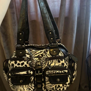 Svartvit axelväska med djurmönster - Cool axelväska från angelkiss i svart och vitt med leopard- och zebramönster. Väskan har flera fickor med dragkedja och snygga detaljer i svart skinnimitation. Perfekt för dig som gillar att sticka ut med din stil.