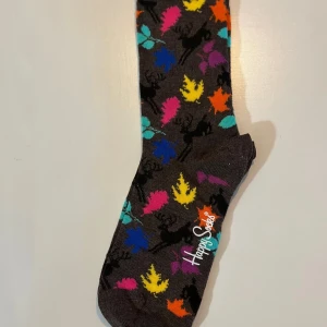Färgglada strumpor från Happy Socks strl 36-40 - Mörkbruna strumpor från Happy Socks med färgglada löv i blått, gult, rosa, orange, turkost och lila. Strumporna har ett lekfullt mönster och är tillverkade i mjuk bomullsmix. Perfekta för att liva upp outfiten.