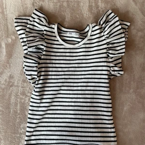 Randig ribbad topp med volangärm Zara - Superfin ribbad topp från Zara i svart och vitt med horisontella ränder. Toppen har korta volangärmar som ger en extra touch och rund halsringning. Perfekt för dig som gillar en trendig och lekfull stil.