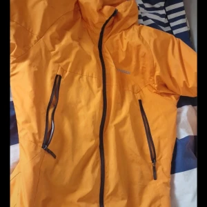 Orange vindjacka från Helly Hansen - Snygg orange vindjacka från Helly Hansen med svarta dragkedjor och hög krage. Jackan har två snedställda fickor med dragkedja och är perfekt för blåsiga dagar. Materialet är lätt och smidigt, och jackan har en sportig vibe.