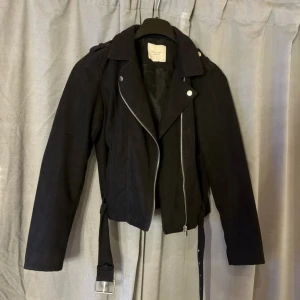 Svart bikerjacka från Jacqueline de Yong - Svart bikerjacka från Jacqueline de Yong i storlek 36. Jackan har klassisk sned dragkedja, långa ärmar, axelklaffar och bälte i midjan. Snygg och stilren modell i bomullsmaterial, perfekt för dig som gillar en edgy look.