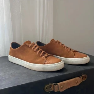 Bruna sneakers från Sneaky Steve - Snygga bruna sneakers från Sneaky Steve med ovandel i mocka och detaljer i skinn. Skorna har vit platt sula och klassisk snörning. Perfekta för dig som gillar stilrena och tidlösa sneakers med en twist av premiumkänsla.