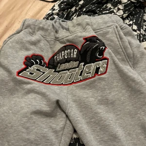 Trapstar grå mjukisbyxor London Shooters - Grå mjukisbyxor från Trapstar med stort broderat London Shooters-motiv och svart panter på framsidan. Byxorna har snörning i midjan och dragkedja vid benslut. Sköna och stilrena, perfekta för en streetwear-look. Passar om man är från 165 och uppåt. Priset kan diskuteras vid snabbaffär 