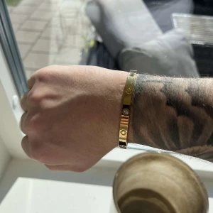 Guldigt armband med stenar - Snyggt armband i guldig metall med infällda klara stenar och små ingraverade symboler runt om. Stilren och modern design som passar perfekt för att lyfta din outfit. Armbandet har en slät, polerad yta och är lätt att matcha med andra accessoarer. Oanvänd 
