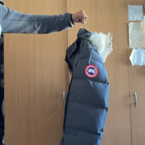 Grå Canada Goose dunjacka med päls - Säljer en grå dunjacka från Canada Goose med ikonisk patch på ärmen och fluffig päls runt huvan. Jackan har dragkedja och knappar framtill, stora fickor och en klassisk pufferlook. Perfekt för kalla vinterdagar och riktigt snygg streetstil. Tags och dustbag ingår!