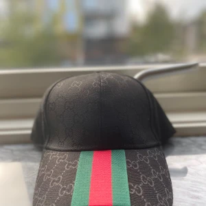 Svart Gucci keps med GG-mönster - Snygg svart keps från Gucci med diskret GG-monogram över hela tyget och klassiska gröna och röda ränder framtill. Justerbart spänne i guld med Gucci-logga baktill. Tillverkad i Italien och i bomullsmaterial. Perfekt för dig som vill ha en lyxig och trendig accessoar.