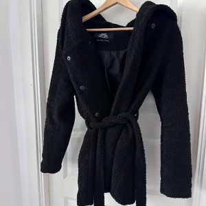 Svart teddyjacka med huva Viva Couture - Mysig svart teddyjacka från Viva Couture med stor huva och knytbälte i midjan. Jackan har knappar framtill och är fodrad på insidan för extra komfort. Perfekt för dig som vill ha en varm och trendig look under kyliga dagar.